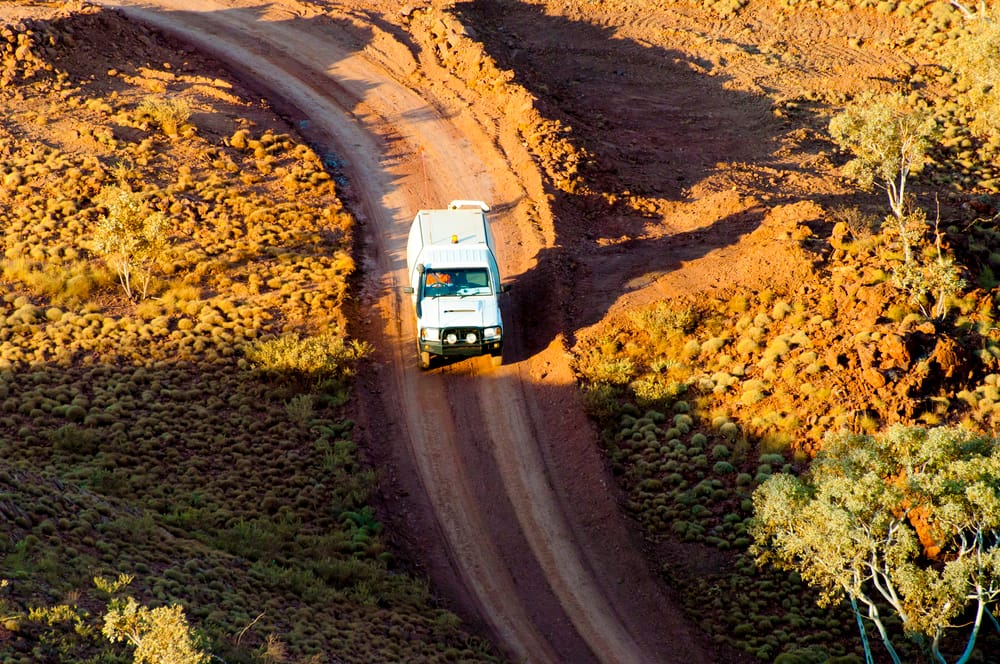 Aerial view of a white 4WD with a rooftop pack driving a wide red-dirt track through Australian outback